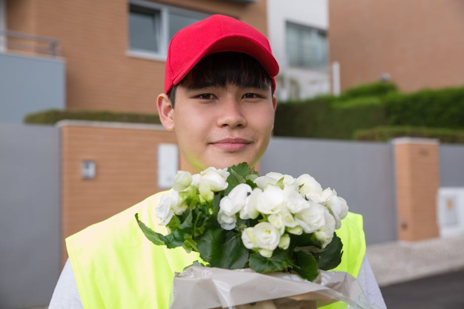 A young male flower delivery courier wearing a bright yellow high-visibility vest and red cap is holding a bouquet of white roses wrapped in light paper with green leaves visible. He stands outdoors i