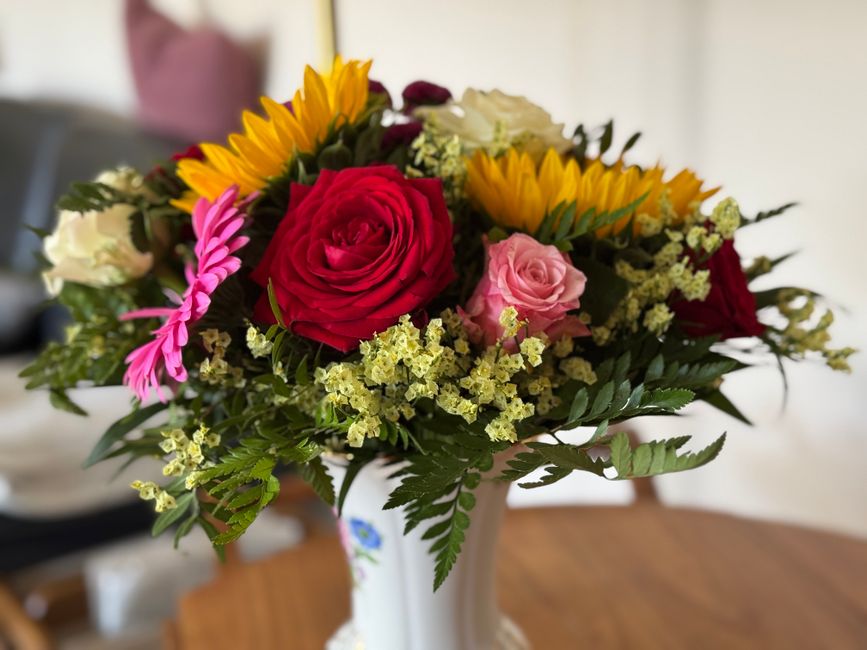A vibrant floral bouquet arranged in a white ceramic vase featuring a combination of fresh roses in shades of red, pink, and white, complemented by yellow sunflowers, pink daisy-like flowers, and deli