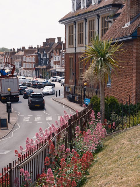 A vibrant floral display along a sloped grassy area in Esher, featuring pink and red astilbe flowers and a tall, spiky palm tree, with a black metal fence separating the garden from a busy street. Beh