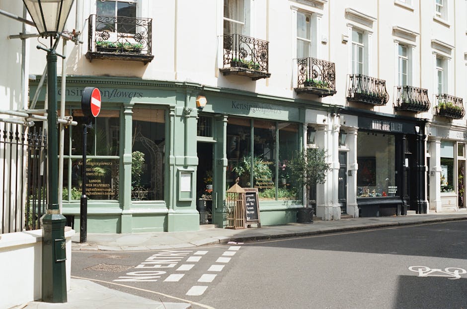 A storefront with large glass windows displaying floral arrangements and greenery inside, painted in a soft pastel green color, located on a street corner with a black chalkboard sign outside. The bui