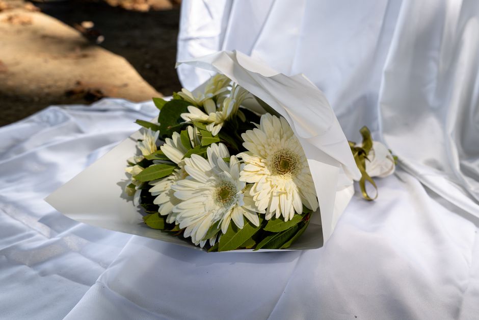 A bouquet of fresh white gerbera daisies with bright yellow centers, paired with green foliage, wrapped in elegant white paper and tied with a golden ribbon, resting on a white satin cloth, with soft 