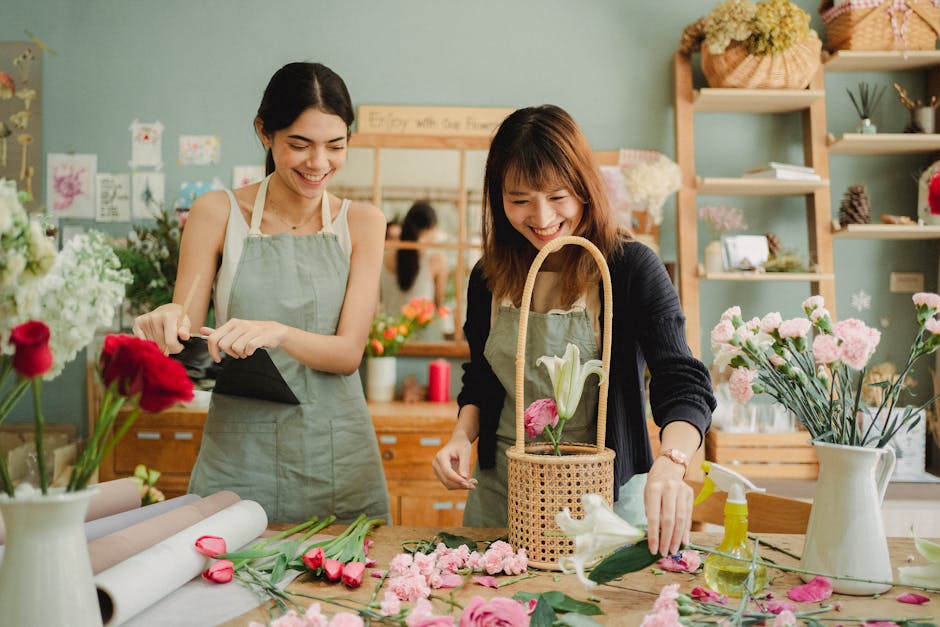 Two women in a florist shop are arranging a floral gift consisting of white lilies and pink carnations, surrounded by various fresh flowers including pink roses and pink carnations on a wooden table. 