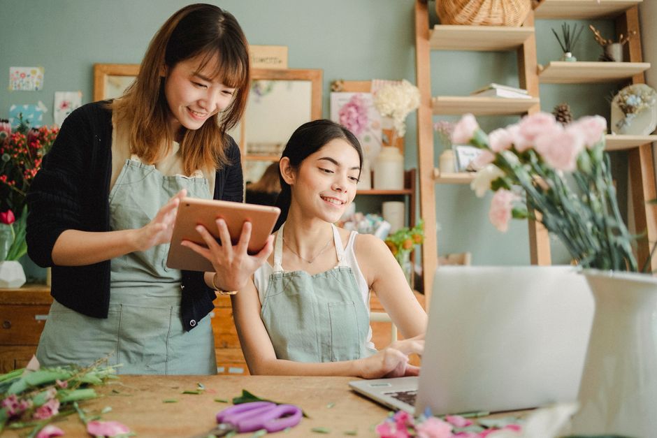 A smiling woman with dark hair in a white top and light green apron sits at a wooden table inside a flower shop, surrounded by fresh flowers and floral materials. A second woman with shoulder-length b